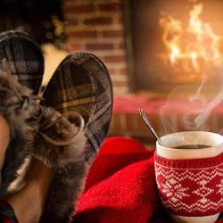 Cozy scene featuring feet in fuzzy slippers resting on a red blanket, with a steaming mug by a crackling fireplace. Warm ambiance invites relaxation.