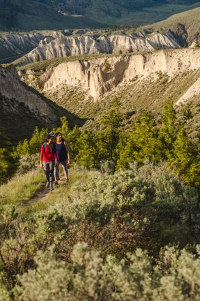 Two hikers traverse a winding trail through lush greenery, with rugged cliffs and rolling hills in the background, showcasing a scenic landscape.