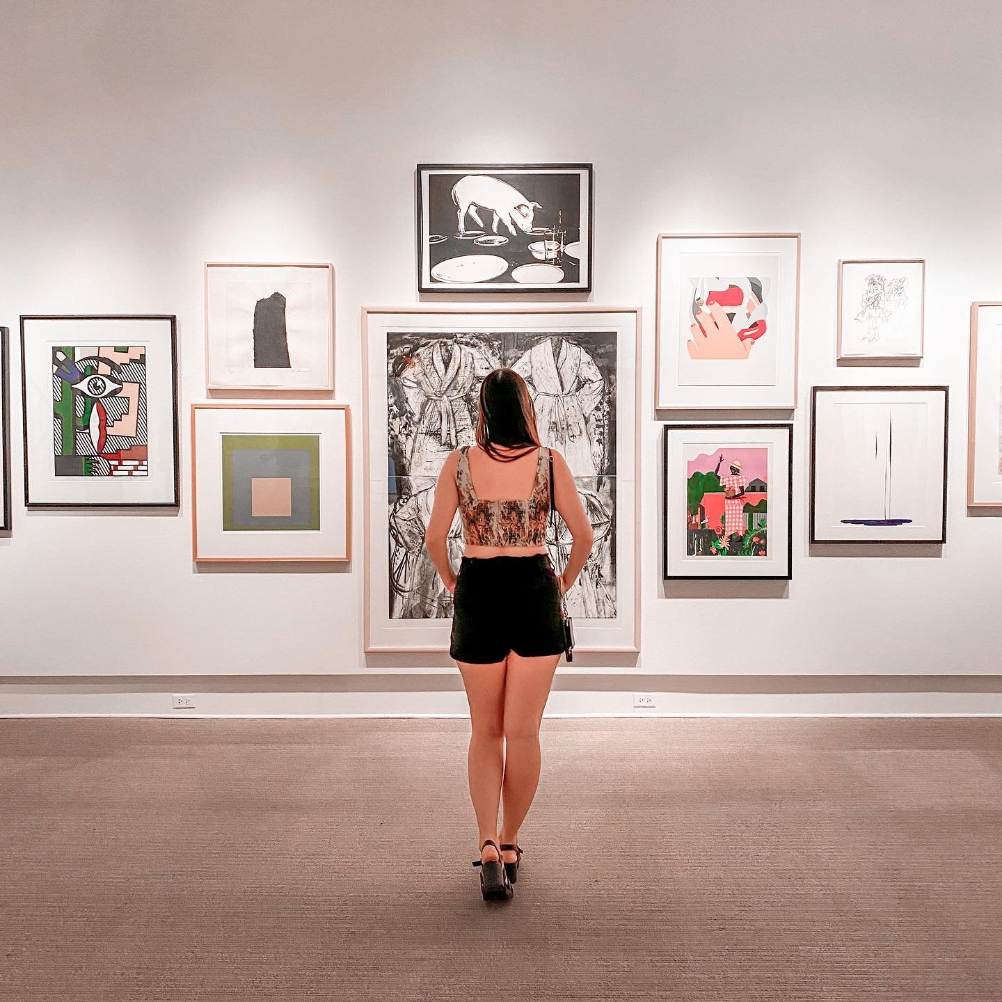 A visitor stands in an art gallery, observing various framed artworks on the walls, showcasing diverse styles and colors. The space is well-lit and inviting.