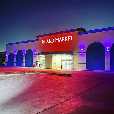 Exterior view of Island Market at night, featuring illuminated signage and colorful lighting. The building has arched entrances and a spacious parking area.
