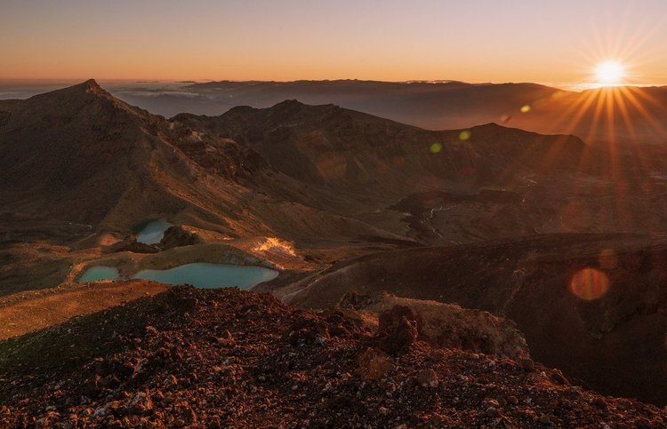 Photo by littlerobinphotographynz, caption reads: A ridiculously early morning had us hiking up to greet the sun on the Tongariro Alpine Crossing 🥾

We hiked under the full moon without the need to use head torches, reaching the high point overlooking Emerald Lakes before the sun rose over the far horizon 🌄

Sunbeams bathed the mountains and valleys in soft golden light, painting the volcanic terrain red. A cool wind whipped around us, freezing out fingers, but we didn't care - we were completely in awe of this otherworldly dreamscape that seemed too perfect for human eyes ✨