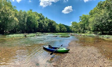 A green and blue kayak rests on a rocky riverbank, surrounded by lush green trees and a clear blue sky with fluffy clouds. The calm water reflects the vibrant scenery.