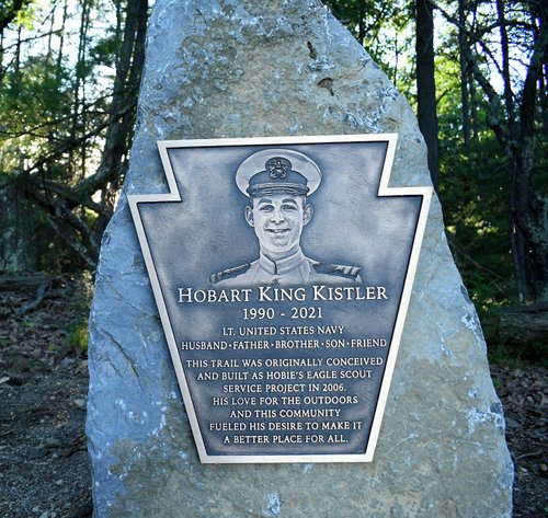 A stone memorial plaque featuring a portrait and text honoring Hobart King Kistler, surrounded by trees in a serene outdoor setting.