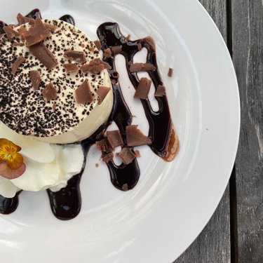 A creamy dessert topped with chocolate shavings and drizzled with chocolate sauce, served on a white plate with a flower garnish.