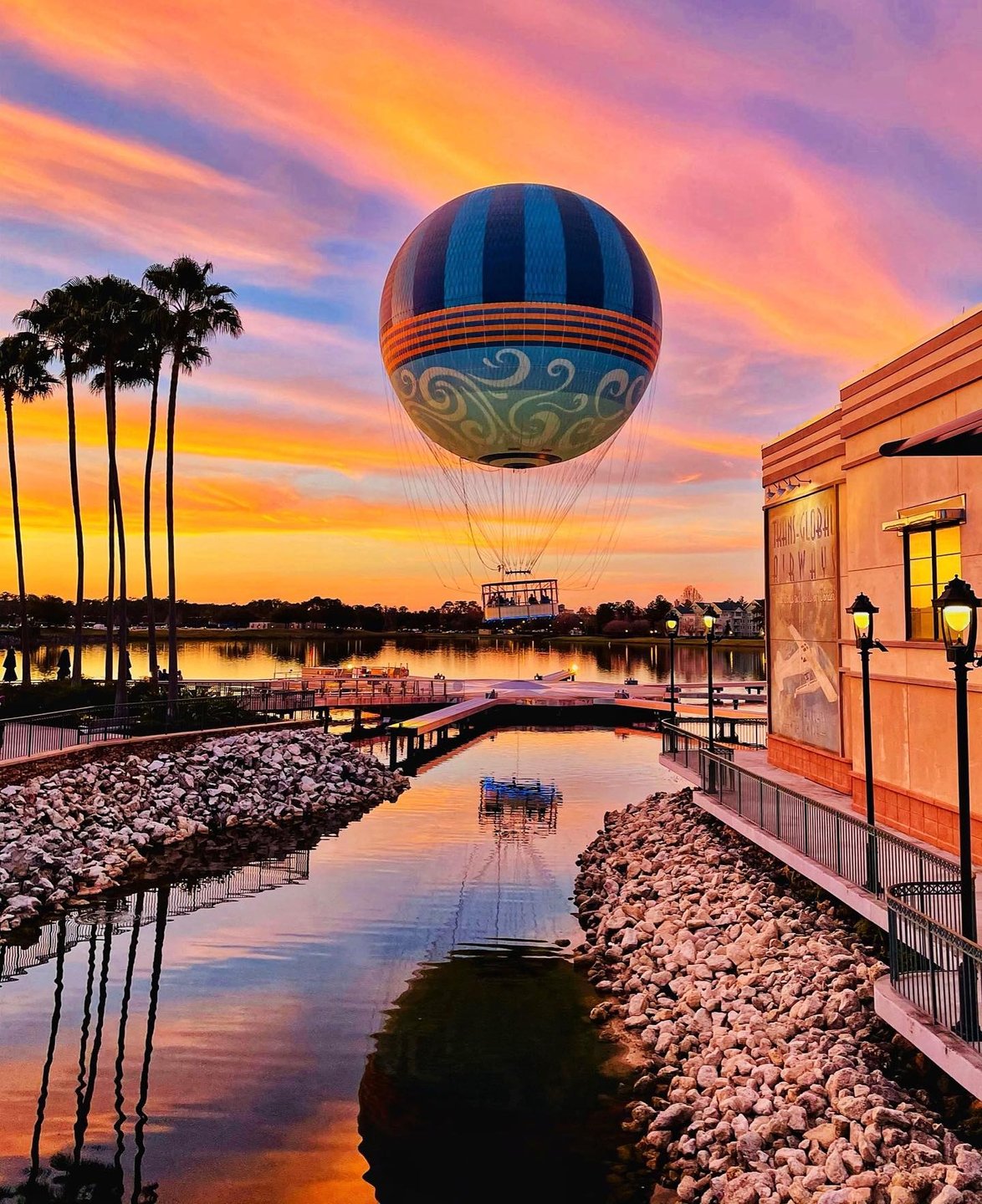 A colorful hot air balloon floats above a serene lake at sunset, reflecting vibrant hues in the water, surrounded by palm trees and pathways.
