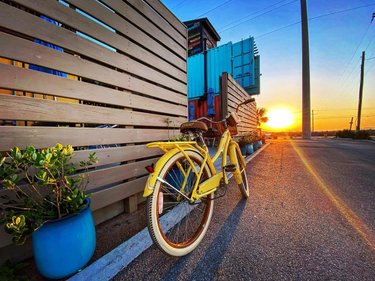 A yellow bicycle leans against a wooden fence at sunset, with colorful containers in the background and potted plants nearby.