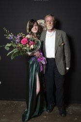 A woman in a green gown holds a bouquet of flowers, standing next to a man in a suit. They pose against a dark backdrop.