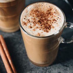 A close-up of a frothy coffee drink topped with cinnamon, served in a clear glass mug, with cinnamon sticks nearby on a dark surface.