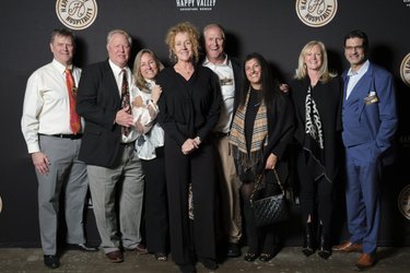 Group of seven people posing together in front of a dark backdrop with "Happy Valley" branding, smiling and dressed in formal attire.