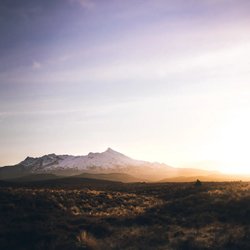 Sunset over a vast landscape with rolling hills and a snow-capped mountain range in the distance, casting warm golden light across the terrain.