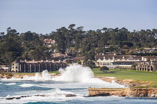 Coastal view featuring crashing waves against rocky shore, with a luxury resort and lush trees in the background, promoting a serene getaway.
