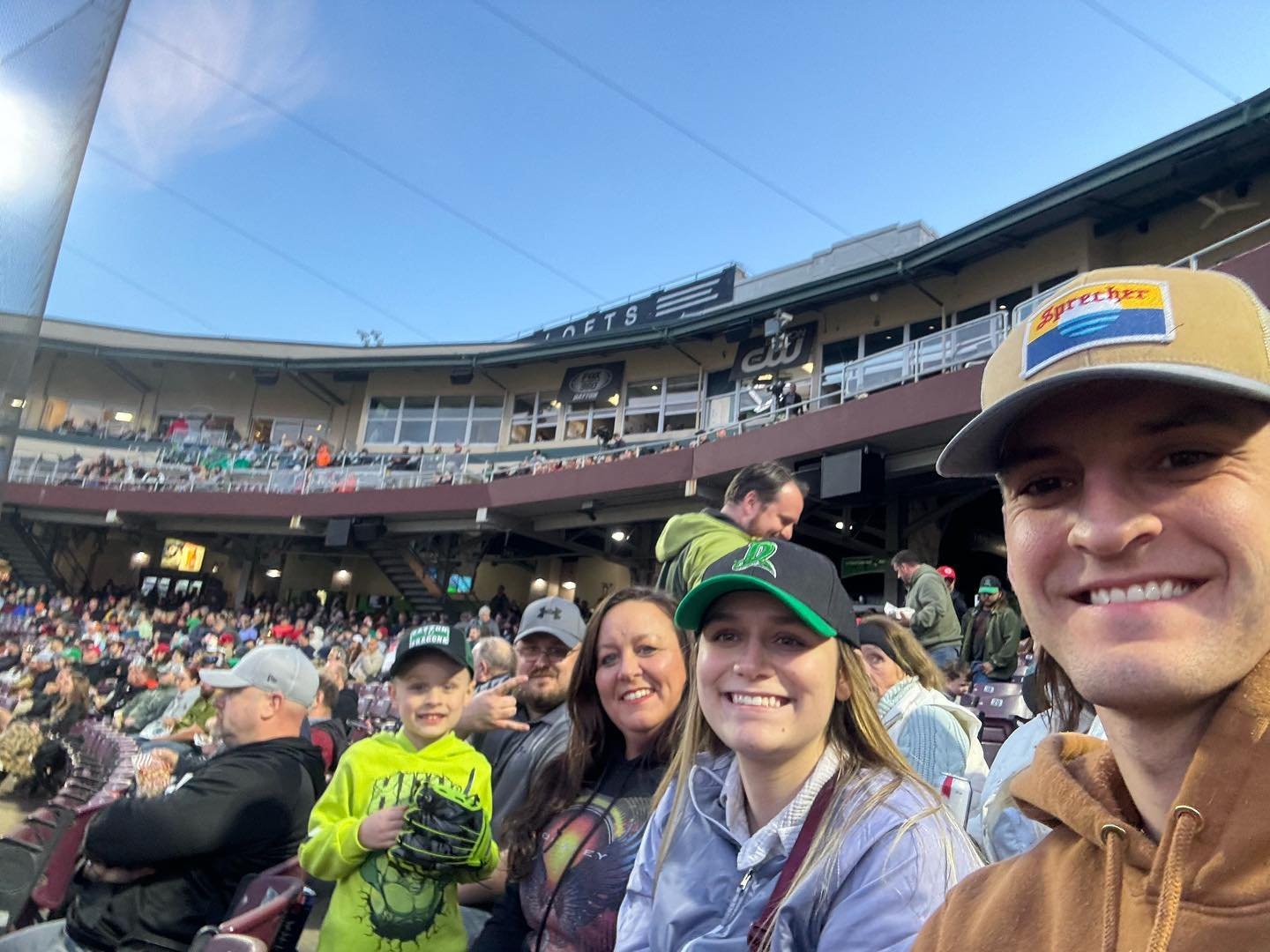 A group of five people, including a child, smiles for a selfie at a lively sports event. The stadium…
