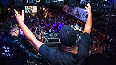 A DJ performs on stage with hands raised, surrounded by a lively crowd in a dimly lit venue, creating an energetic atmosphere.