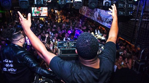 A DJ performs on stage with hands raised, surrounded by a lively crowd in a dimly lit venue, creating an energetic atmosphere.