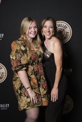 Two women pose together in front of a dark backdrop featuring a circular logo. One wears a floral dress, while the other is in a sleek black outfit.