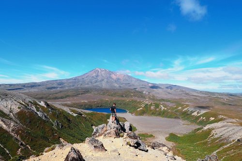 A hiker stands on rocky terrain overlooking a vast landscape with a lake and a prominent mountain in the background under a clear blue sky.