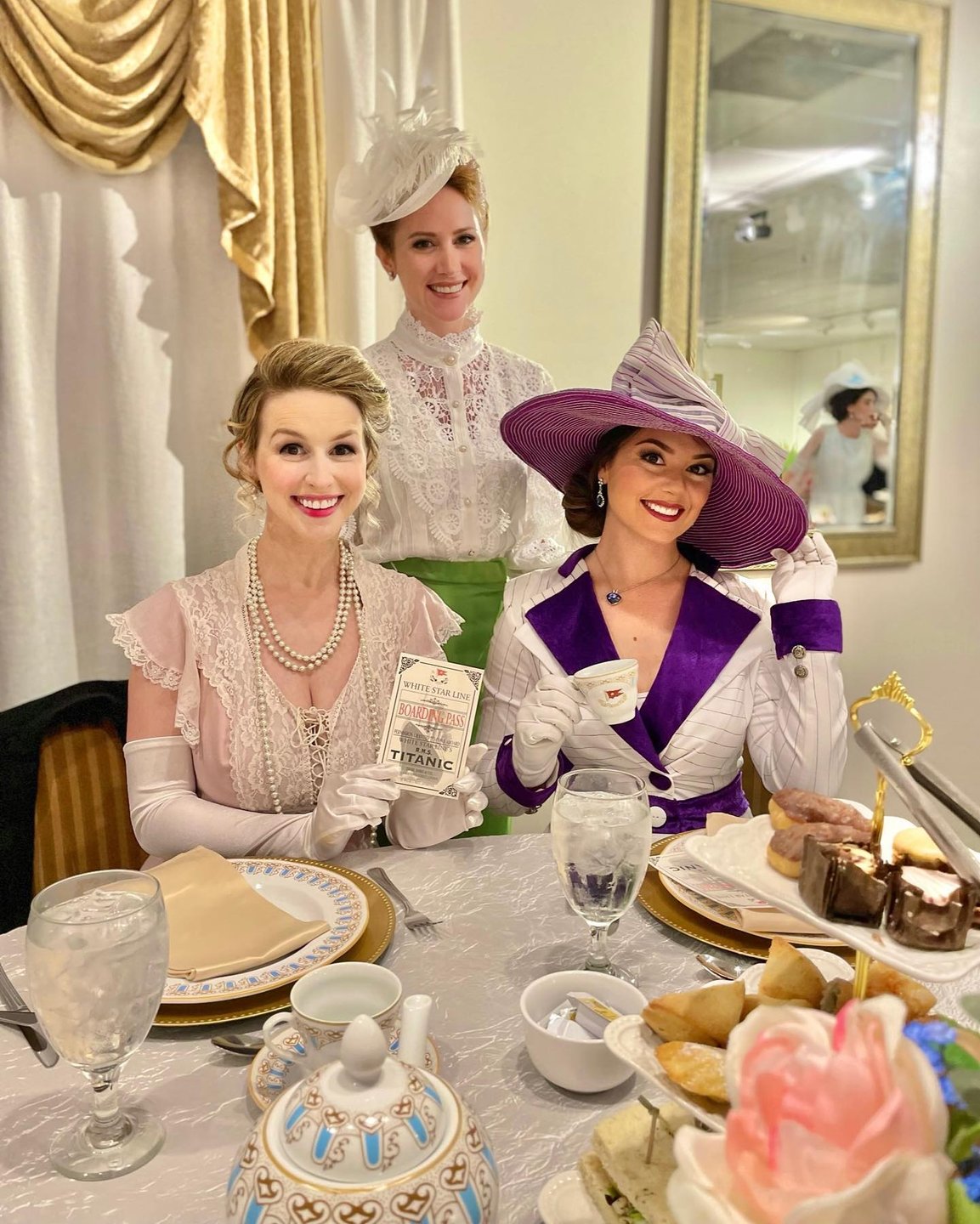 Three elegantly dressed women sit at a beautifully set table, enjoying a tea service with fine china, pastries, and decorative flowers.