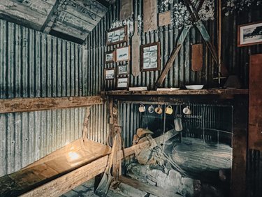 Interior of a rustic cabin featuring corrugated metal walls, wooden furniture, and framed photographs. A stone fireplace is visible.