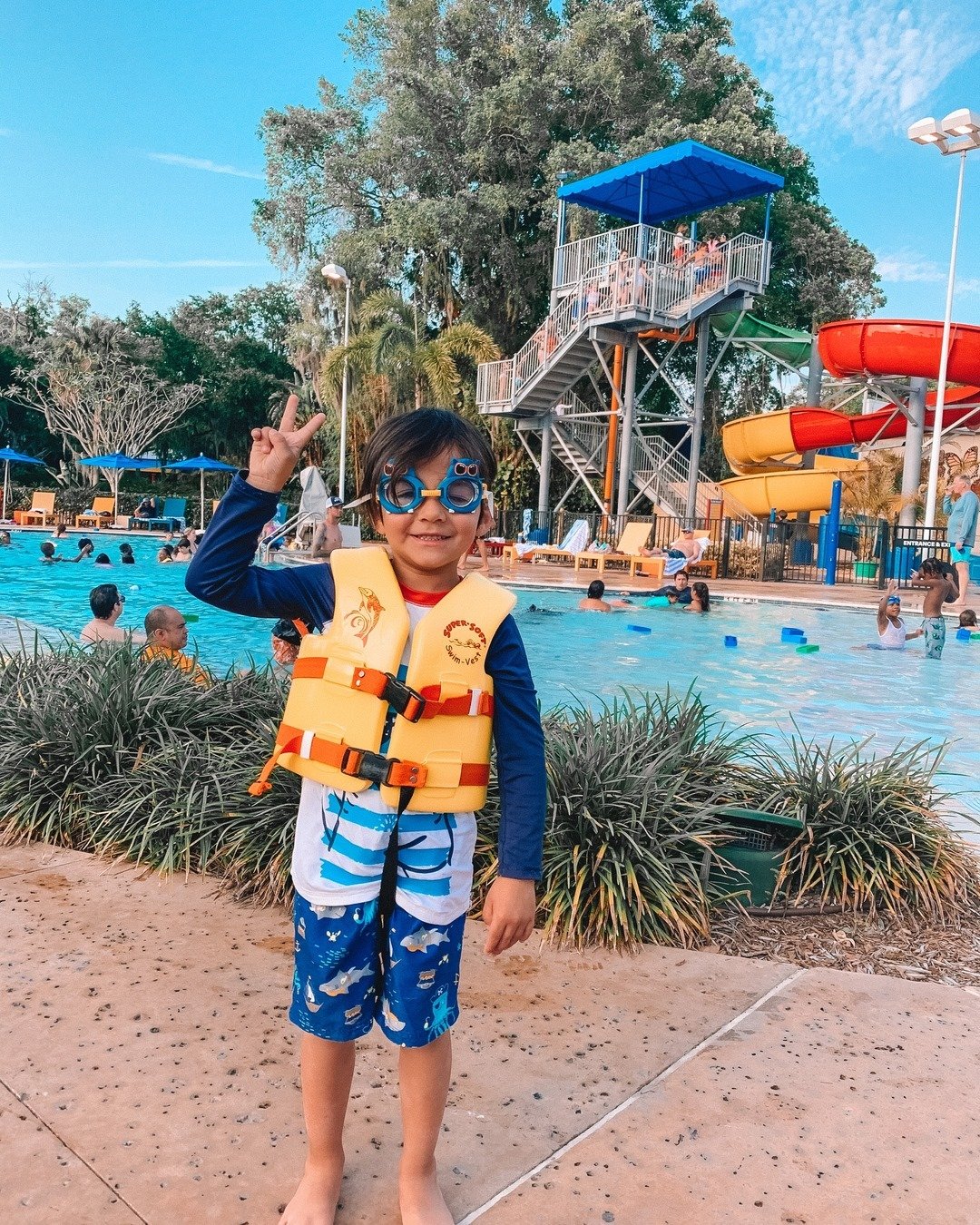 A child in a life jacket and goggles poses by a vibrant water park pool, featuring slides and playful water features in the background.
