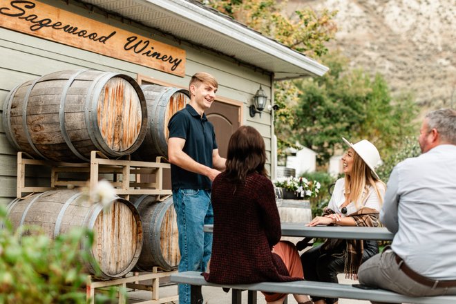 A young man engages with three people at a picnic table outside Sagewood Winery, surrounded by wine barrels and greenery.