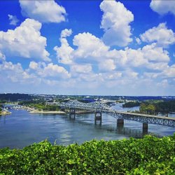 A scenic view of a river with a bridge spanning across it, surrounded by lush greenery and dotted with boats. Fluffy clouds fill the blue sky.