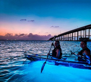 Two women in illuminated kayaks paddle on calm waters at dusk, with a bridge in the background and a colorful sunset sky.