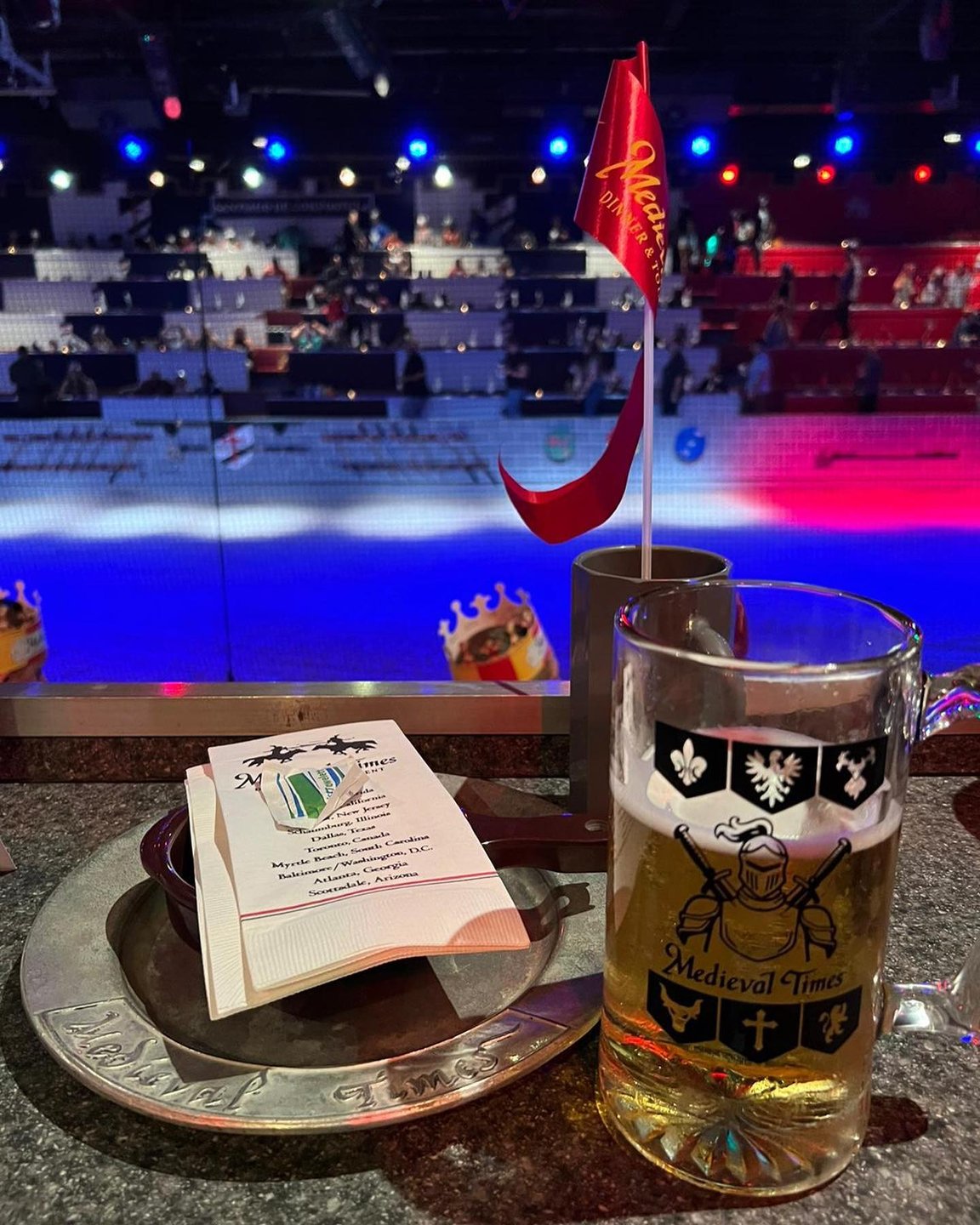 A beer mug on a silver tray with a menu, overlooking a brightly lit arena with colorful lights and empty seating. A flag is visible.