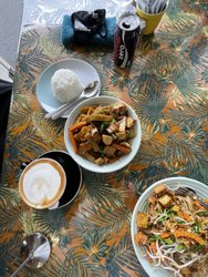 A vibrant table setting featuring a bowl of stir-fried vegetables, rice, and a plate of noodles, alongside a coffee and a soda.