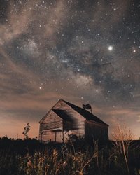 A weathered barn stands under a starry night sky filled with the Milky Way, surrounded by tall grass and distant trees.